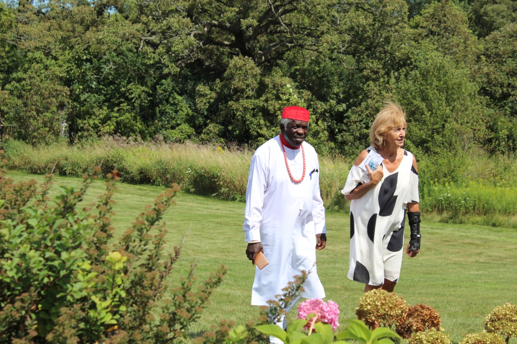 The bride’s father, a Nigerian in traditional Igbo garb, walking in a meadow with the groom’s aunt, a white woman in a black and white dress. 