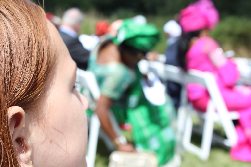 A red-headed, ten-year-old girl’s face is close-up in the left-hand corner. In the blurred background, across the aisle seated in chairs for a wedding are women in traditional Igbo dress, bright green and bright pink. 