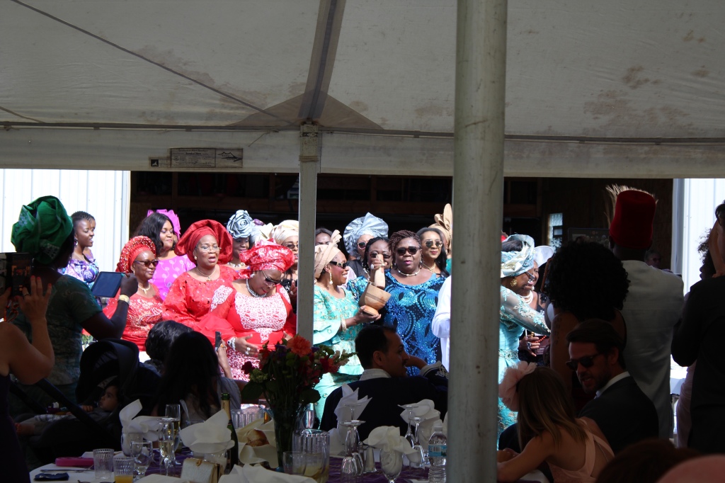 Taken from the back of an outdoor tent, there is a group of women in Igbo dress dancing outside the tent. They are doing a dance to indicate their acceptance of the groom’s family for their daughter/cousin—this shows they will not take her away and nullify the wedding. In the foreground are spectators, other wedding guests witnessing the transaction. 