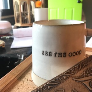 Stoneware coffee mug that says "See the good" with the edge of a leather journal in the foreground
